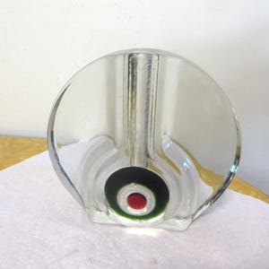 May include: Clear glass vase with a circular design. The vase has a central cylindrical opening and a decorative element with a red dot surrounded by green and black circles. The vase has a rounded shape.