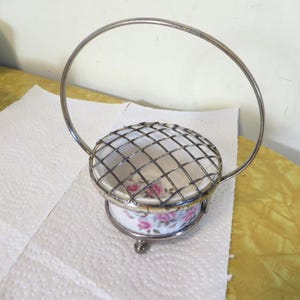 May include: A silver wire basket with a handle and a white ceramic base with a floral design. The base has a wire mesh top.