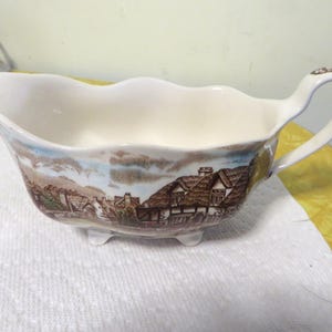 May include: A white ceramic gravy boat with a brown and blue cottage scene design. The boat has a scalloped edge and three feet.