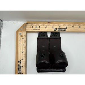 May include: A dark brown leather double pouch with two snap closures, positioned against a wooden ruler showing a width of approximately 3.5 inches and a height of approximately 5 inches. The pouch features two vertical straps with metal snaps.