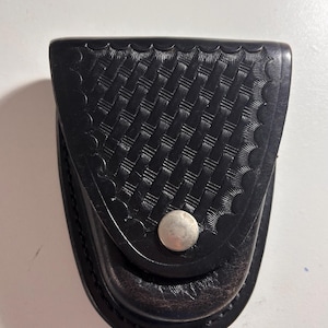 May include: Black leather handcuff case with a basketweave pattern on the front flap. The case has a scalloped edge and a silver snap closure. The bottom of the case is a darker shade of black.