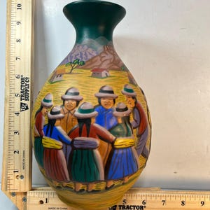 Może przedstawiać: Ręcznie malowany ceramiczny wazon z ciemnozieloną szyjką i szeroką podstawą. Wazon przedstawia scenę ludzi w tradycyjnych strojach, z żółtym tłem i krajobrazem gór i domów. Wazon ma około 25 cm wysokości.