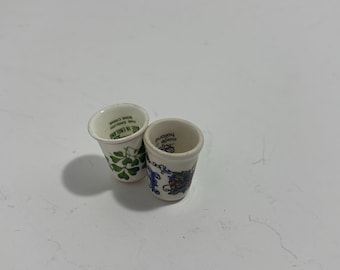 Könnte beinhalten: Zwei kleine, weiße Keramik-Fingerhüte. Einer hat ein grünes Kleeblattmuster, der andere ein blaues und rotes Blumenmuster mit dem Text "made in Holland". Beide sind zylindrisch mit leicht ausgestelltem Oberteil.