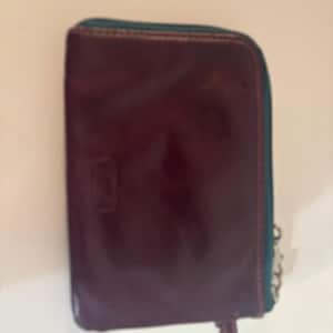 Puede incluir: Una cartera rectangular de cuero burdeos oscuro con cremallera y ribete turquesa. La cartera tiene un pequeño diseño en relieve en la parte delantera y una cadena plateada sujeta a la cremallera. El cuero tiene un acabado brillante.