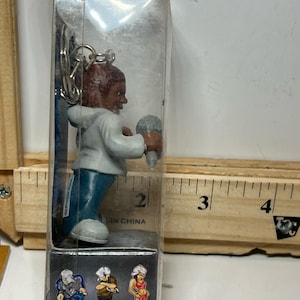 Könnte beinhalten: Miniaturfigur einer Person mit weißem Kapuzenpullover und blauer Jeans, die ein Mikrofon hält. Die Figur ist an einem Schlüsselanhänger befestigt. Die Verpackung enthält drei weitere Miniaturfiguren. Das Produkt befindet sich in einer transparenten Kunststoffhülle.