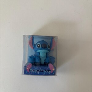 Op de afbeelding: Blauw beeldje van een stripfiguur met grote oren en een brede glimlach, in een doorzichtige plastic doos. Het beeldje heeft roze accenten op de oren en voeten. De basis van de doos heeft het woord "Sacapuntas".