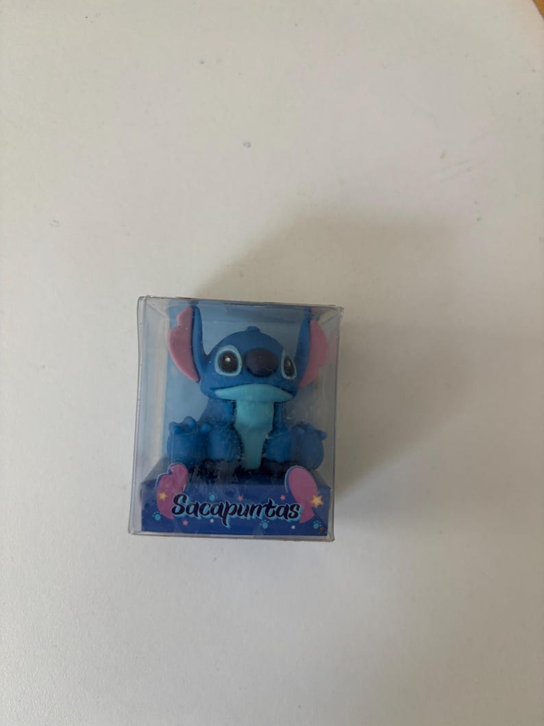 Op de afbeelding: Blauw beeldje van een stripfiguur met grote oren en een brede glimlach, in een doorzichtige plastic doos. Het beeldje heeft roze accenten op de oren en voeten. De basis van de doos heeft het woord "Sacapuntas".