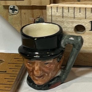 May include: A miniature ceramic mug shaped like a man's face wearing a black top hat. The mug has a white interior and a gray handle. The face is painted with realistic skin tones and facial features.