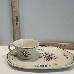 May include: Cream-colored teacup and saucer set with floral designs in red, green, and blue. The cup has a handle and sits on a matching oblong saucer with a green border. A wooden ruler is in the background.