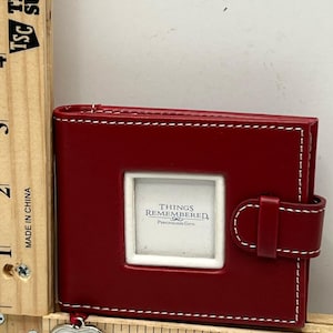 Puede incluir: Un álbum de fotos de cuero rojo con costuras blancas y un marco rectangular para una foto. El álbum tiene un cierre de hebilla y un dije en forma de corazón que dice "DIANE". El texto "THINGS REMEMBERED" es visible.