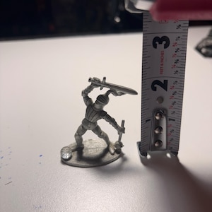May include: A silver-colored miniature knight figurine, approximately 2.5 inches tall, wielding a sword and a mace. The knight is posed in an action stance, with a helmet and armor. The figurine is on a round base.
