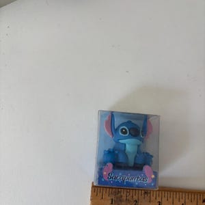 Op de afbeelding: Een blauw Stitch-beeldje in een doorzichtige plastic doos. Het beeldje heeft grote oren en &eacute;&eacute;n oog. Op de doos staat het woord "Sacapuntas". Een houten liniaal ligt naast de doos.