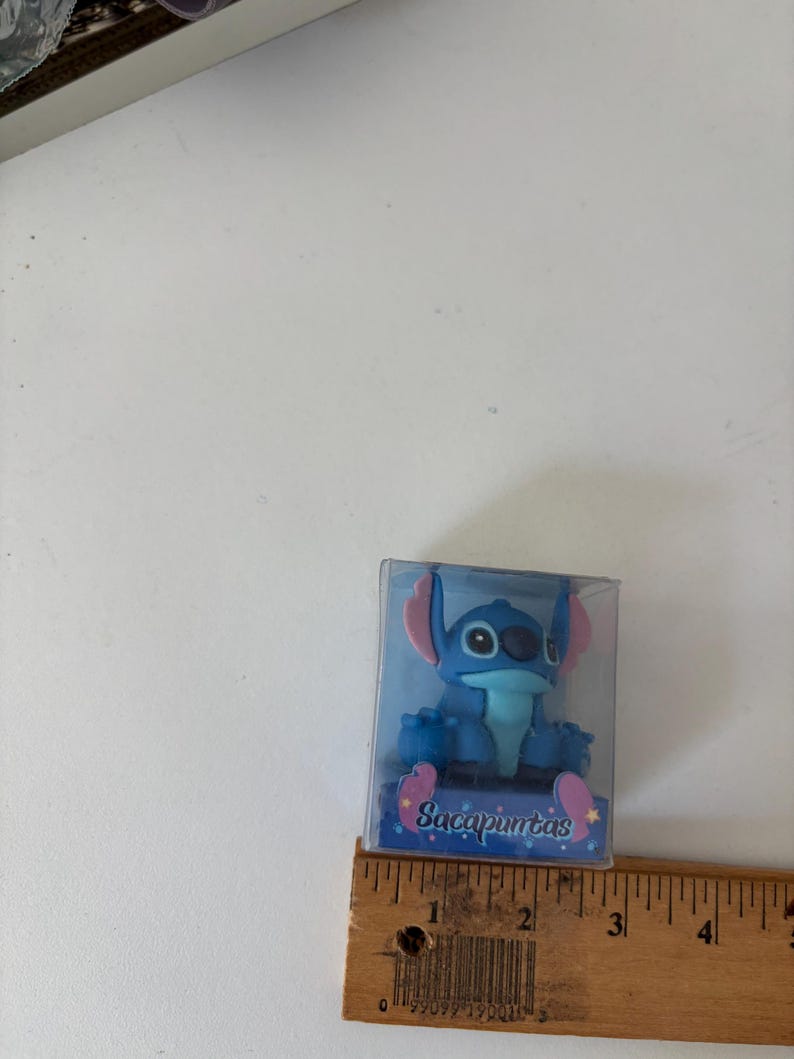 Op de afbeelding: Een blauw Stitch-beeldje in een doorzichtige plastic doos. Het beeldje heeft grote oren en &eacute;&eacute;n oog. Op de doos staat het woord "Sacapuntas". Een houten liniaal ligt naast de doos.