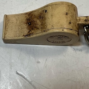 May include: A vintage off-white whistle with a metal ring. The whistle has dark stains and a circular embossed design on the bottom. The whistle is made of plastic and has a curved shape.