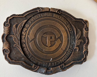 Könnte beinhalten: Eine Vintage-Gürtelschnalle in Kupferoptik mit dem Text "PASTORE MUSIC 507-3424-5 UNION CITY, IN." Die Schnalle hat ein zentrales kreisförmiges Design mit dem Buchstaben "P" und Pfeilen, umgeben von dekorativen Schnörkeln.