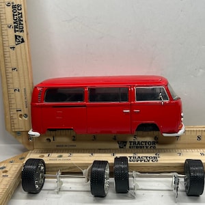 May include: A red toy van with four detached wheels. The van has a classic design with windows and doors. The wheels are black with silver rims. A wooden ruler is placed next to the van, showing measurements in inches.