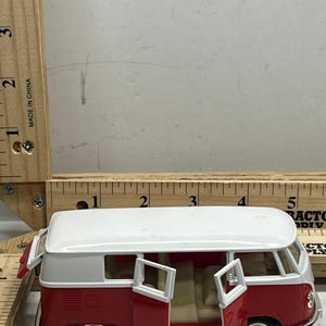May include: A red and white toy van with its doors open, revealing a beige interior. The van has black tires and chrome hubcaps. The toy is next to a wooden ruler with inch measurements.