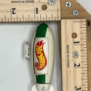 May include: A vintage Hot Wheels toy car in the shape of a rocket, with a white body, green accents, and the Hot Wheels logo. The toy is approximately 3 inches long.