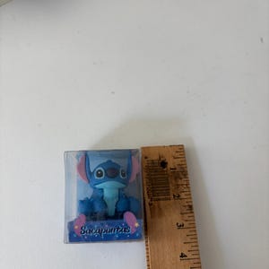Op de afbeelding: Een blauw Stitch-beeldje in een doorzichtige plastic doos. Het beeldje heeft grote oren en een vriendelijke uitdrukking. Op de doos staat het woord "Sacapuntas". Een houten liniaal meet ongeveer 5 cm.