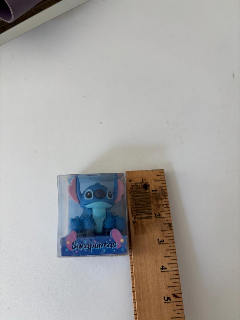 Op de afbeelding: Een blauw Stitch-beeldje in een doorzichtige plastic doos. Het beeldje heeft grote oren en een vriendelijke uitdrukking. Op de doos staat het woord "Sacapuntas". Een houten liniaal meet ongeveer 5 cm.