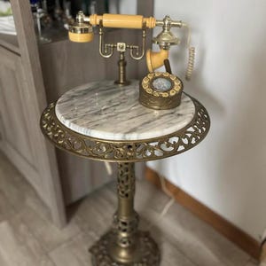 Puede incluir: Un teléfono antiguo y una mesa auxiliar. El teléfono tiene un auricular color crema y un dial giratorio. La mesa tiene una tapa de mármol y detalles de latón ornamentados. La mesa mide aproximadamente 61 cm de altura.