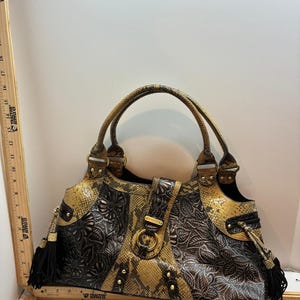 May include: A handbag featuring a combination of yellow and black snakeskin-patterned panels and dark brown embossed floral leather panels. The bag includes two handles, gold-tone hardware, a central strap with a ring, and black tassels on the sides.