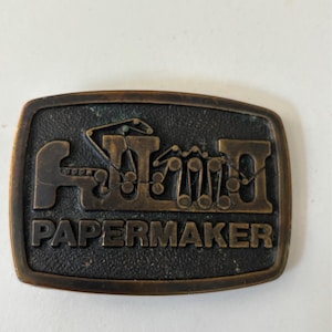 May include: A rectangular, bronze-colored belt buckle with the word "PAPERMAKER" in capital letters. The buckle features a stylized illustration of machinery. The background has a textured appearance.