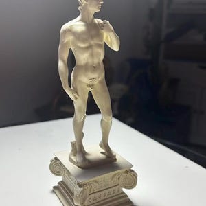 Peut inclure: Statue crème d'une figure masculine nue, debout sur un piédestal carré avec des sculptures ornées. La statue est une représentation classique, avec une musculature détaillée et une expression sereine. Le piédestal porte l'inscription "CAESAR".