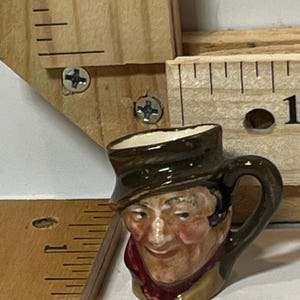 May include: A small, ceramic mug with a handle, featuring a painted face of a man wearing a hat and scarf. The mug is brown with a white interior and is approximately 1 inch tall.