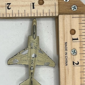 Puede incluir: Un pequeño avión de juguete beige con detalles plateados, colocado cerca de una regla de madera. La regla, marcada en pulgadas, indica que el avión mide aproximadamente 5 cm de largo. Se lee "MADE IN CHINA" en la regla.