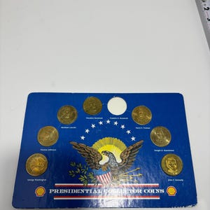 Könnte beinhalten: Eine blaue Sammlungsanzeige mit goldfarbenen Münzen mit Porträts US-amerikanischer Präsidenten. Die Anzeige enthält den Text "Presidential Collector Coins" und das Shell-Logo. Die abgebildeten Präsidenten sind George Washington, Thomas Jefferson, Abraham Lincoln, Theodore Roosevelt, Franklin D. Roosevelt, Harry S. Truman, Dwight D. Eisenhower und John F. Kennedy.