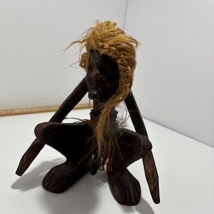 May include: A dark wooden figurine of a person in a seated position. The figure has a braided, light brown hairpiece and is wearing a light brown rope around the neck. The figure is carved with visible facial features and is sitting with its legs bent.