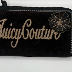 May include: Black Juicy Couture wallet with a zip closure. The wallet features the brand name embroidered in a cursive font and a decorative flower. The zipper pull has the Juicy Couture logo.