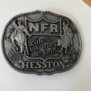 May include: A silver-colored belt buckle with a rodeo theme. The buckle features the text "NFR 25th Anniversary Series" and the name "Hesston." It also depicts a cowboy riding a horse and a bull rider. The buckle has the numbers "59" and "83."