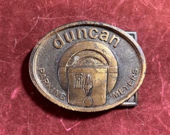 May include: Vintage Duncan Parking Meters belt buckle. The oval buckle is made of bronze-coloured metal. The text "duncan" is at the top, with "PARKING METERS" on the sides. The centre features a parking meter design with the word "EXPIRED".