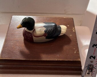 Puede incluir: Una figura decorativa de pato de madera con cabeza negra, pico amarillo y cuerpo blanco, negro y granate. El pato está montado sobre una base de madera marrón oscuro. La base mide aproximadamente 15 cm de largo.