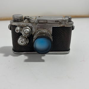 May include: A vintage-style camera figurine with a silver-colored body and a textured, dark brown leather-like grip. The camera features a prominent blue lens and several silver-colored dials and buttons.