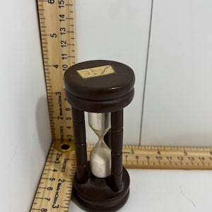 Puede incluir: Un pequeño reloj de arena marrón oscuro con interior plateado. El reloj de arena está hecho de madera y vidrio, y mide aproximadamente 10 cm de alto. La parte superior del reloj de arena tiene una pequeña pegatina redonda.