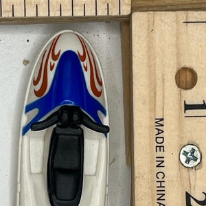 May include: A small, white toy jet ski with a blue and red flame design on top. The jet ski has a black seat and handlebars. The image includes a wooden ruler with inch measurements.