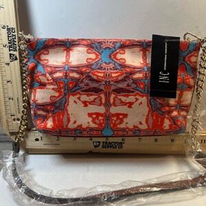 May include: A clutch purse with a colorful abstract pattern in shades of red, blue, and cream. It has a gold chain strap and a brown leather strap. The tag reads "INC INTERNATIONAL CONCEPTS".