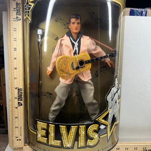 May include: A boxed Elvis Presley doll, labelled "Teen Idol." The doll is dressed in a pink and white checked jacket, grey trousers, and holds a decorated guitar. The box is gold with the name "ELVIS" and the text "THE SUN NEVER SETS ON A LEGEND."