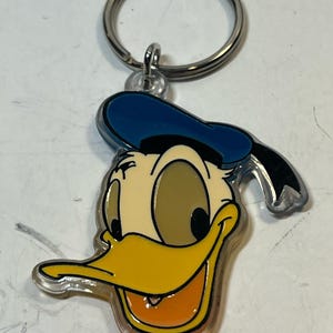 May include: A Donald Duck keychain featuring a smiling face with a yellow beak, orange mouth, and blue hat. The keychain has a silver metal ring for attaching to keys or bags. The design is outlined in black and has a clear plastic border.