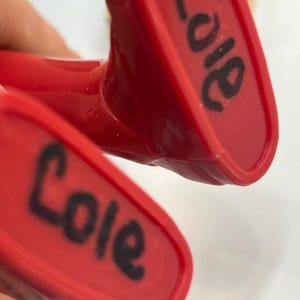 May include: Close-up of two red plastic objects with the name "Cole" written in black marker on each. The objects appear to be part of a larger item, possibly a toy or tool.