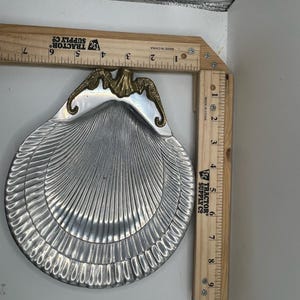 May include: A silver-toned metal shell-shaped dish featuring a gold-toned decorative accent at the top. The dish is positioned against two wooden rulers, showing a width of approximately 7 inches and a height of approximately 9.5 inches.
