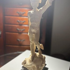 Puede incluir: Una estatua de color crema de una figura de pie, posiblemente un personaje mitológico, sosteniendo un objeto en alto. La figura está sobre una base texturizada, montada sobre un pedestal cuadrado negro. La estatua tiene un diseño detallado y está bien iluminada.