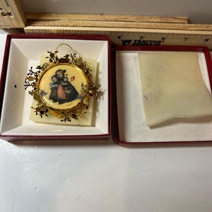 Könnte beinhalten: Runder Weihnachtsschmuck mit einer gemalten Szene von zwei Figuren in Winterkleidung. Der Schmuck ist mit Goldfiligran gerahmt und wird in einer roten Box mit weißem Innenraum präsentiert.