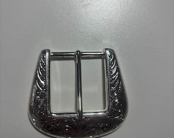 May include: Silver-coloured belt buckle with an ornate, engraved floral design. The buckle has a rectangular opening with a central bar and a curved, decorative outer edge. The metal has a polished finish.