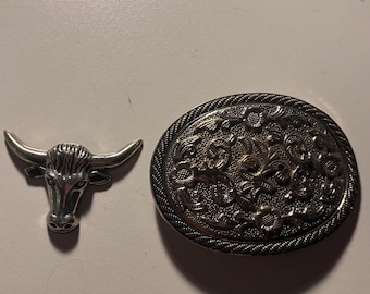 Op de afbeelding: Twee zilverkleurige metalen gespen. Eén is een ovale gesp met een sierlijk bloemmotief en een gevlochten rand. De andere is een stierenkopspeld met hoorns.