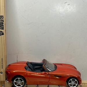 May include: A bright orange convertible toy car with black interior. The car has silver wheels and is positioned on a white surface. The car is approximately 8 inches long.