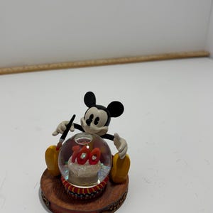 May include: A Mickey Mouse snow globe figurine. Mickey is black, white, and yellow, holding a paintbrush. Inside the globe is a cake with the number 100. The base is brown with the text "100 YEARS OF MAGIC".